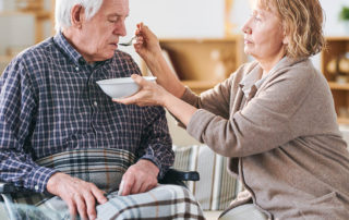 Aged casual woman holding bowl with soup and spoon by her disable husband mouth while helping him to eat dinner